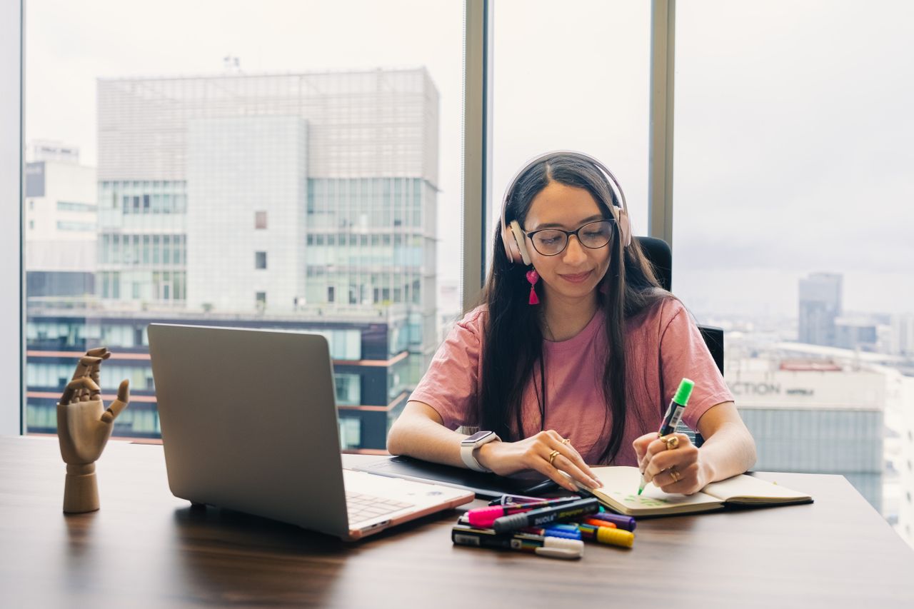 Young woman wearing headphones, writing in notebook at office desk with city view.