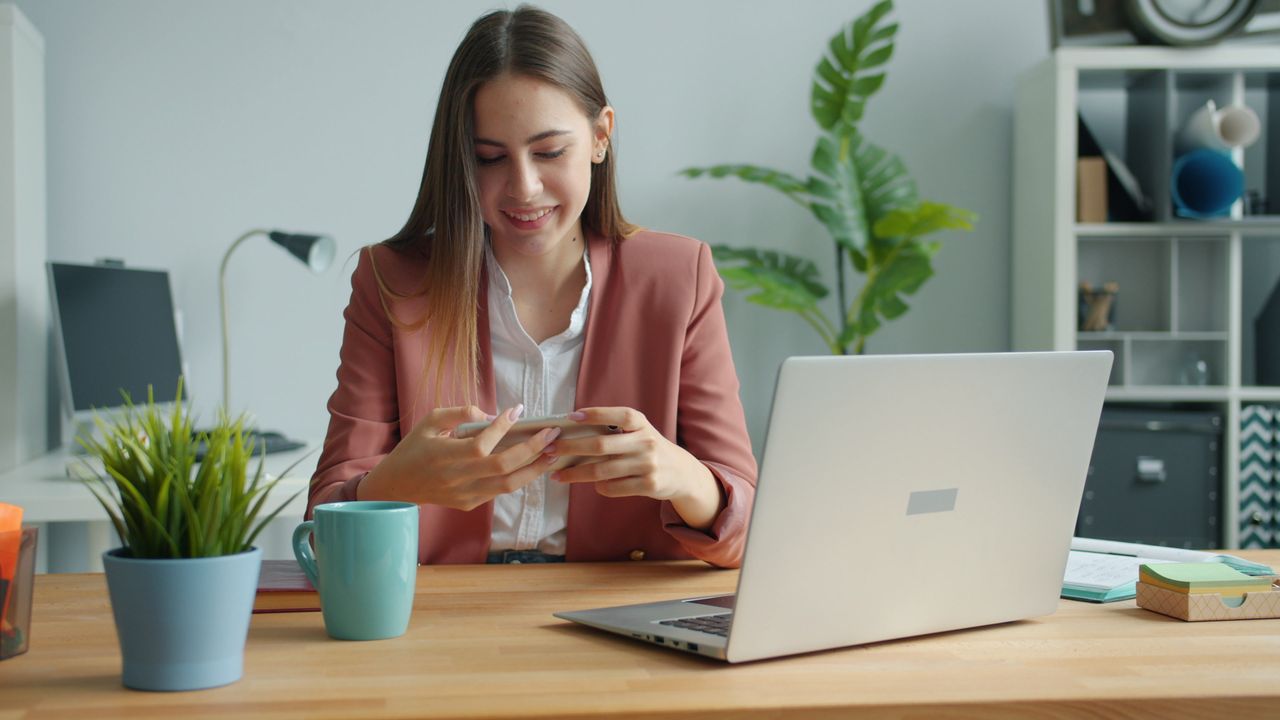Smiling young woman using smartphone at home office desk with laptop and plants.