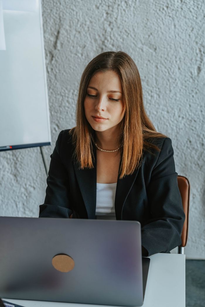 Woman in a black blazer working at a laptop in an indoor office setting.